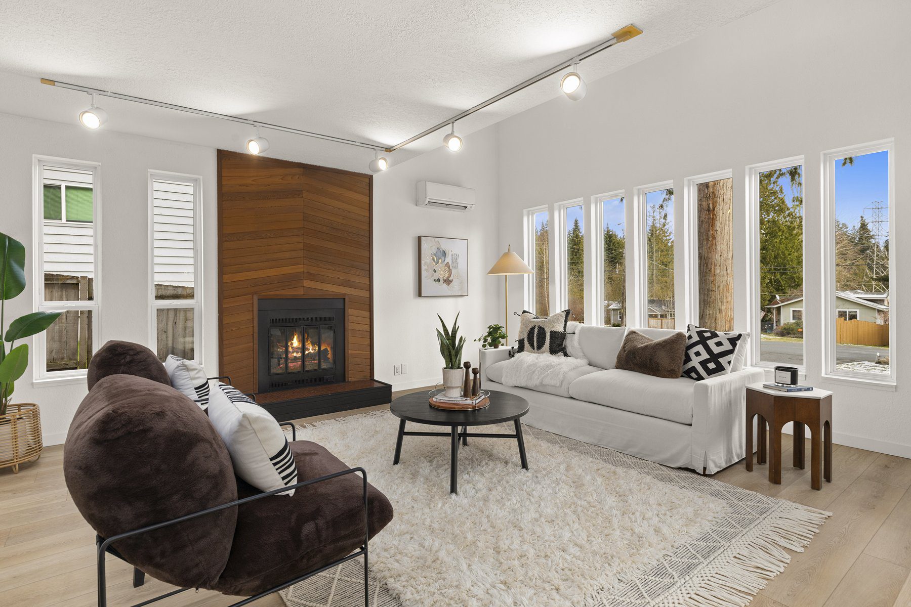 Modern living room with fireplace, white sofa, armchair, round coffee table, large windows, and light wood floors. Neutral decor and ceiling track lighting complete the space.